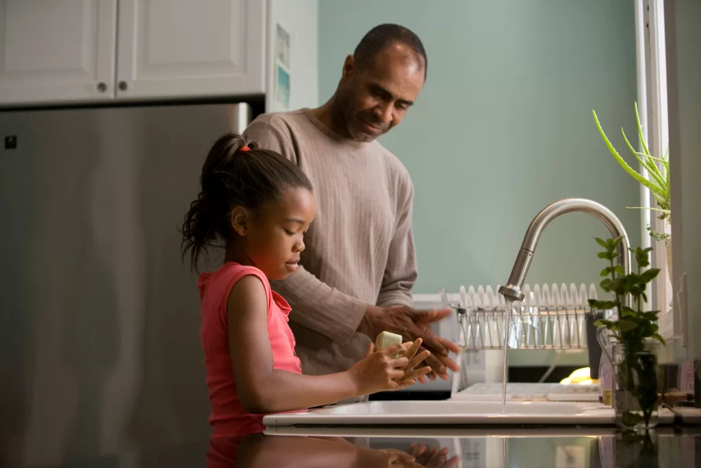 A father watching his young daughter wash her hands with a bar of soap, over a kitchen sink