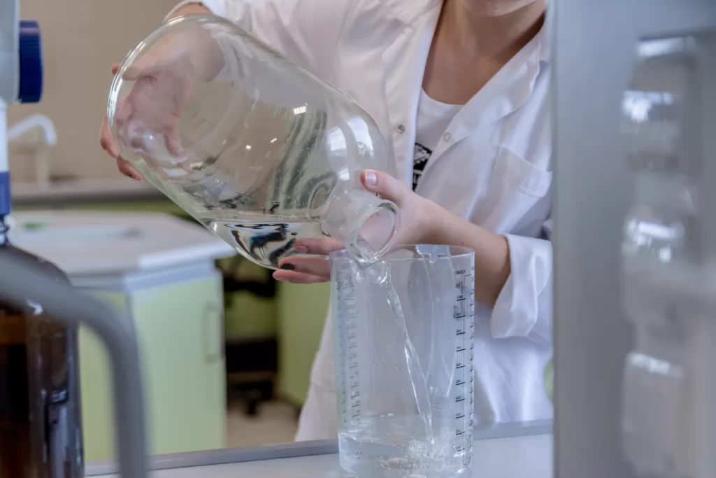  A scientist in a lab coat pouring water from a large, clear glass container into a large beaker
